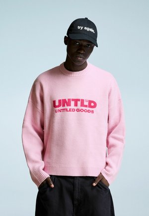Jonge man in een roze trui met de tekst "UNTLD UNTITLED GOODS" en een zwarte pet met de tekst "try again." staat met zijn handen in zijn zakken.