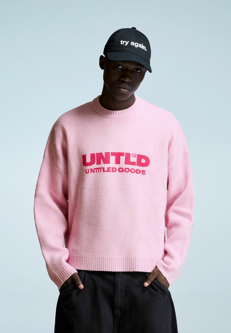 Mlad moški nosi roza pulover z napisom "UNTLD UNTITLED GOODS" in črno kapo z napisom "preizkusi znova." stoji z rokami v žepih.