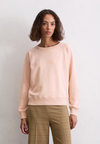 Vaaleanpunainen sweatshirts, jossa on pyöreä kaula-aukko ja raglan-hihat. Edessä on brodeerattu brändin nimi, yhdistettynä ruudullisiin housuihin.