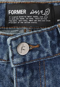 Denim jeans with a faded blue wash, contrasting orange stitching, and a metal button featuring an "F" logo. Label displays brand name "FORMER."