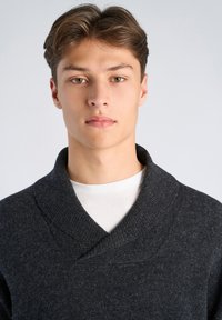 Dunkelgrauer Strickpullover mit Schalkragen, strukturierter Stoff, getragen über einem weißen T-Shirt. Fokus auf Halsausschnitt und Strickdetail. Neutraler Hintergrund.