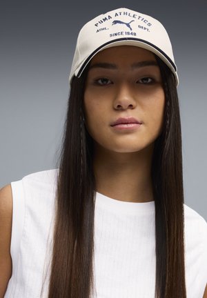 Joven con cabello largo y liso, lleva una gorra beige de Puma Athletics y una camiseta blanca sin mangas, sobre un fondo gris.