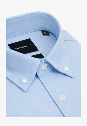 Jasnoniebieska koszula na guziki z kołnierzem zakończonym szpicem, białymi guzikami i czarną metką z logo wewnątrz kołnierza.