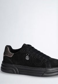 Black suede sneaker with a textured black rubber sole, black laces, and a metallic accent at the heel. Features a pendant logo detail.