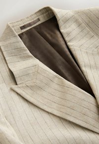 Blazer beige à fines rayures avec de larges revers et une doublure intérieure soyeuse marron foncé, posé à plat sur une surface claire.