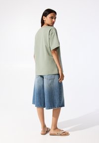 T-shirt vert clair surdimensionné, short en denim bleu délavé à coupe ample et sandales plates beige avec des embellissements. Tenue simple et décontractée.