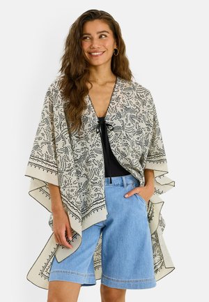 Femme portant un châle beige avec des motifs de feuilles noires, un haut noir, un short en jean bleu clair, souriante, avec une main dans la poche.