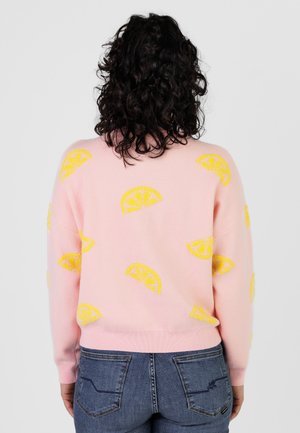 Frau mit lockigem, dunklem Haar trägt einen rosa Pullover mit gelben Zitronenscheibenmustern, kombiniert mit blauen Jeans, Blick von hinten.