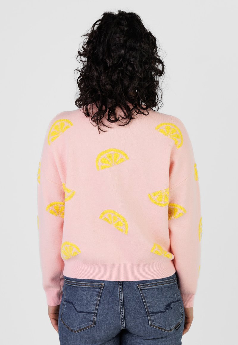 Femme aux cheveux bouclés et foncés portant un pull rose orné de motifs de tranches de citron jaunes, associée à un jean en denim bleu, vue de dos.