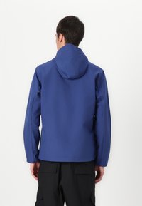 Blå vandtæt jakke med hætte, der har en glat overflade og enkelt design. Synlige lommer på siderne, båret over sorte cargo bukser.