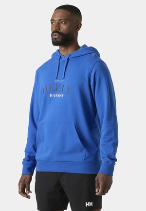 Mężczyzna w jasnoniebieskiej bluzie z kapturem Helly Hansen i czarnych szortach, stojący na tle jasnego, gładkiego tła, zwrócony lekko w lewo.