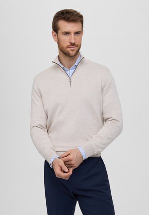 Jersey pullover beige con cierre en el cuello y mangas largas, textura suave, y una camisa de cuello azul claro debajo. Unos pantalones azul marino completan el conjunto.