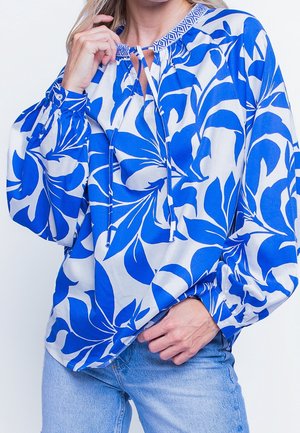 Kvinde iført løs hvid bluse med stort blåt blomsterprint og snøre ved halsudskæringen, kombineret med lyseblå jeans.