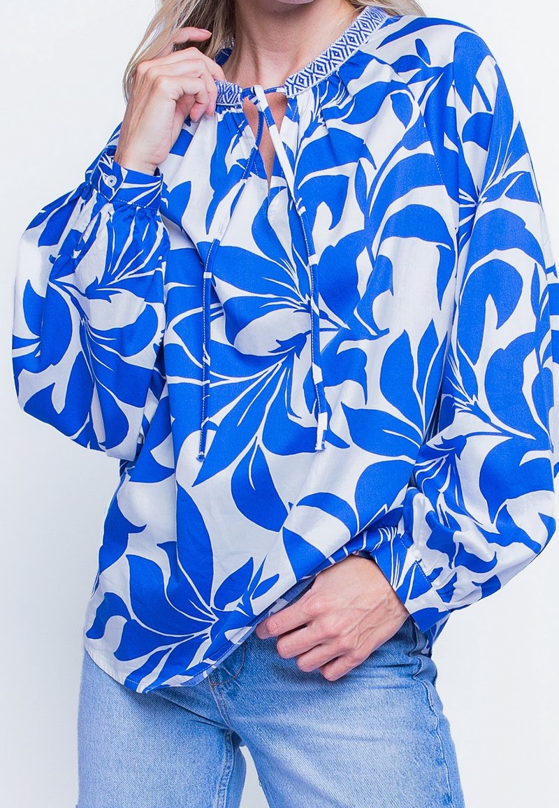 Femme portant un chemisier blanc ample avec un grand imprimé floral bleu et un col à cordon, associé à un jean bleu clair.