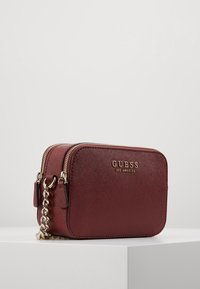 Kompakte burgunderfarbene Leder-Crossbody-Tasche mit goldener Kettenriemen und "GUESS Los Angeles"-Logo auf der Vorderseite, auf einer weißen Fläche platziert.