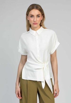 Femme portant une chemise blanche à manches courtes boutonnée, nouée à la taille, et un pantalon large vert olive, se tenant devant un fond uni.
