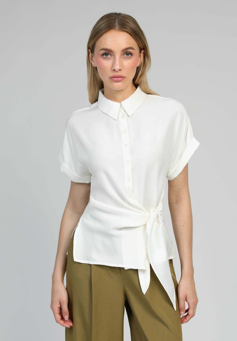 Femme portant une chemise blanche à manches courtes boutonnée, nouée à la taille, et un pantalon large vert olive, se tenant devant un fond uni.
