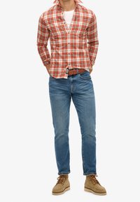 Camicia a quadri rossa e bianca con bottoni, jeans azzurri, stivali beige con lacci. Cintura marrone con fibbia d'argento, vestibilità e stile casual.