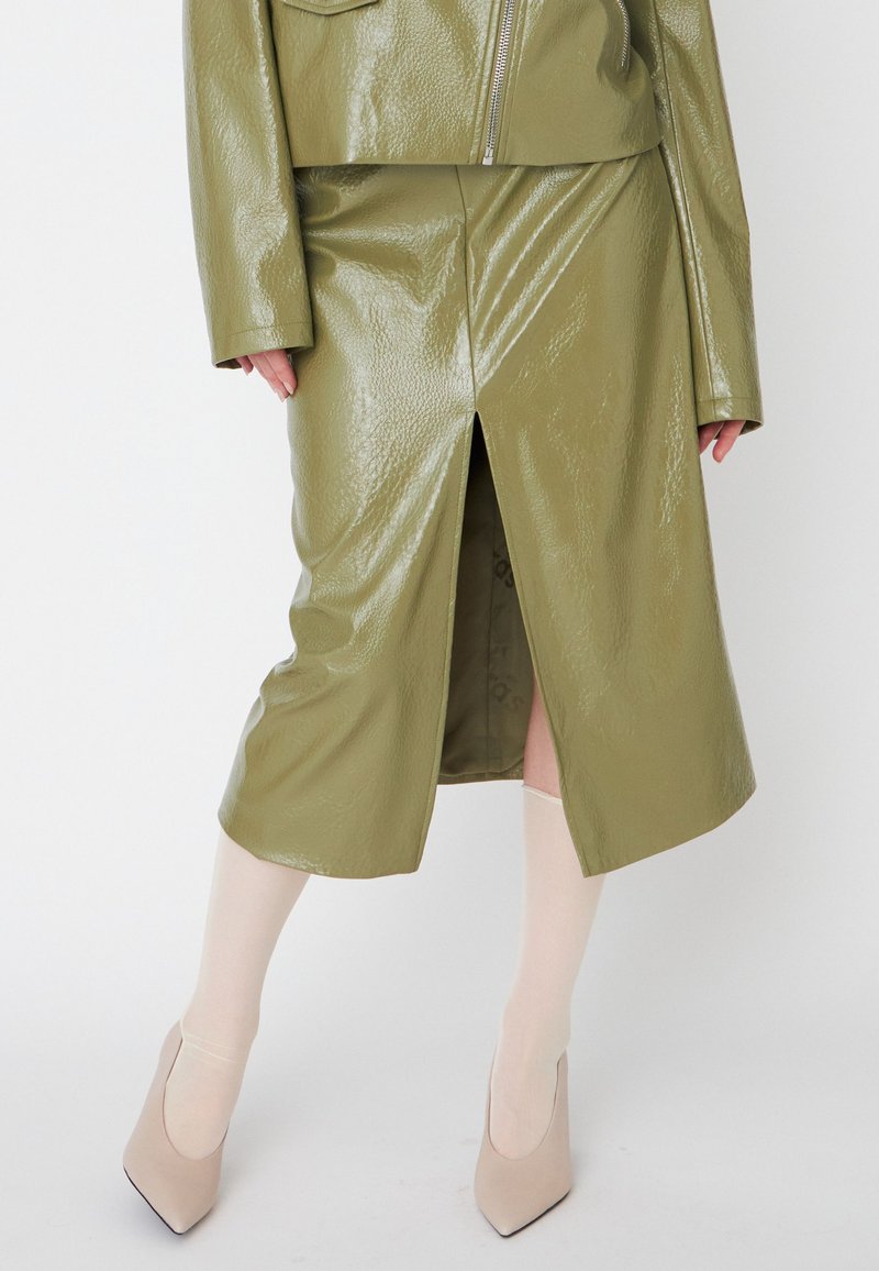 Jupe midi en cuir verni vert olive avec une fente sur le devant et une texture lisse. Assortie à des talons aiguilles beiges et des collants transparents.