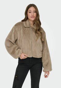 Trumpos zip-up striukės iš minkšto smėlio spalvos dirbtinio kailio su apykakle, pasižyminčios per dideliais rankovėmis ir tekstūruota, pūkuota apdaila. Juodi nusidėvėję džinsai.
