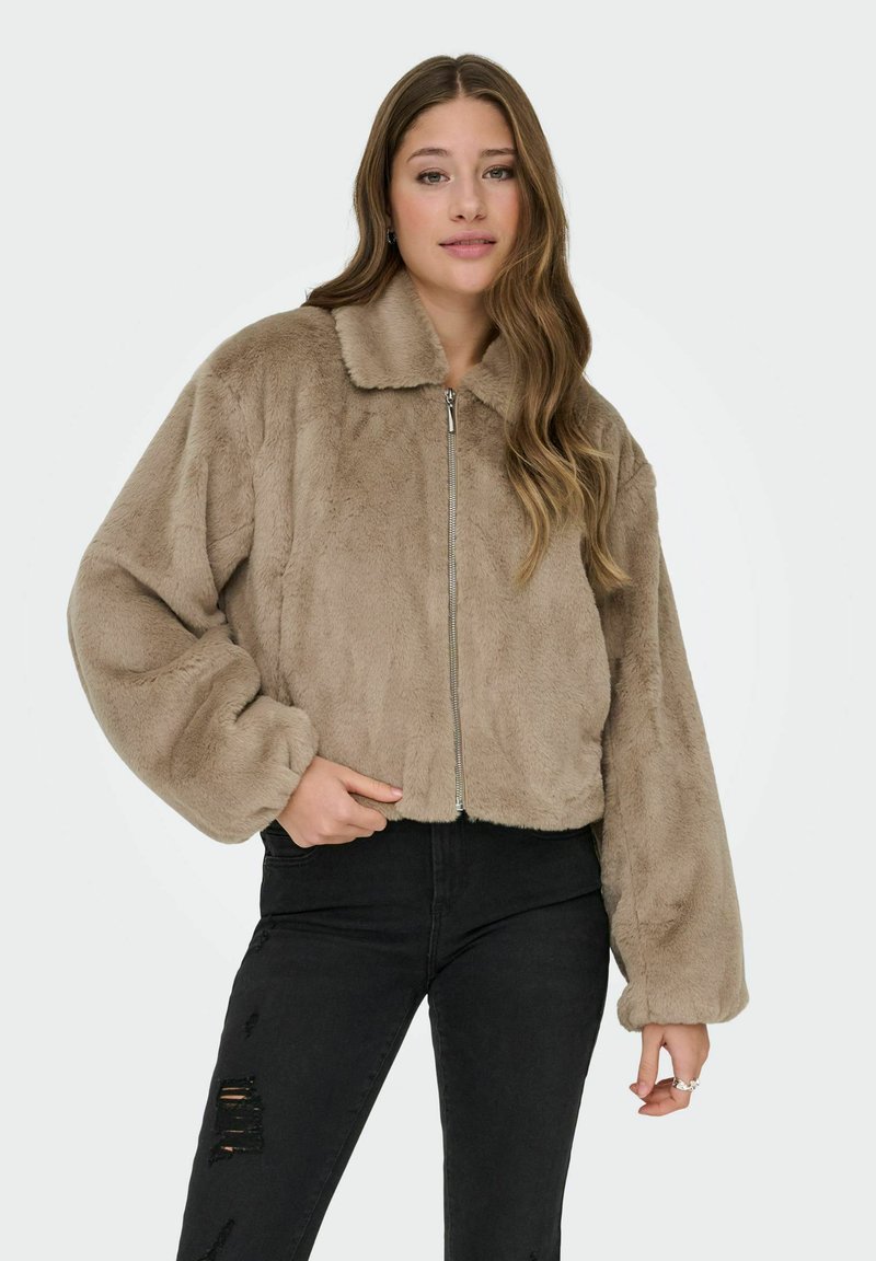 Trumpos zip-up striukės iš minkšto smėlio spalvos dirbtinio kailio su apykakle, pasižyminčios per dideliais rankovėmis ir tekstūruota, pūkuota apdaila. Juodi nusidėvėję džinsai.