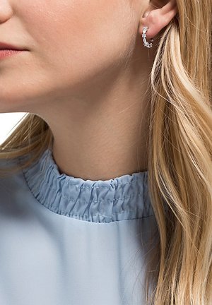 Silver earrings with a wavy design, featuring small clear gemstone accents, worn on the lobe beside a light blue ruffled collar.