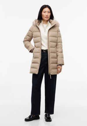 Femme portant un manteau matelassé beige avec une capuche bordée de fourrure, un pull blanc à fermeture éclair, un pantalon large foncé et des mocassins noirs, debout avec une main dans la poche.