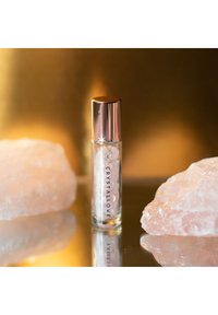 Przezroczysta szklana buteleczka z rolką wypełniona małymi białymi kryształkami, z metalową pokrywką w kolorze różowego złota i etykietą "CRYSTALLOVE", na odbijającej powierzchni.