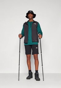 Mann i turutstyr med bøttehatt, grønn langermet skjorte, svart vest og shorts, som holder vandrestaver og står på ensfarget bakgrunn.