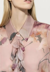 Transparent floral blouse in soft pink with a collar. Features a ruffled front and subtle color variations in the floral pattern. Textured fabric.