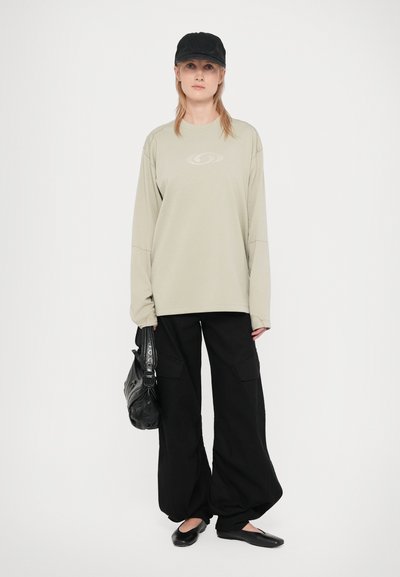 Cămașă verde deschis cu mânecă lungă și un model texturat, pantaloni cargo negri cu picioare largi, pantofi negri fără toc și o șapcă neagră. Geantă neagră.