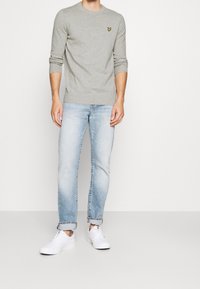 Grijze gebreide trui met lange mouwen en geribbelde manchetten, voorzien van een klein geel vlinderlogo; gecombineerd met lichtblauwe spijkerbroeken en witte sneakers.