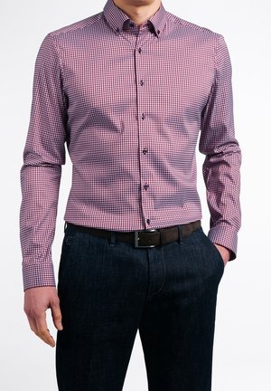 Uomo che indossa una camicia a maniche lunghe con bottoni a quadri rossi e bianchi, infilata in un paio di jeans blu scuro con cintura nera.