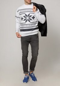 Bílá pletená mikina s černými geometrickými vzory, v kombinaci s tmavými džínami a modrými sportovními botami, držící černou bundu.