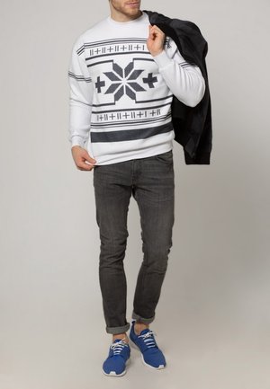 Felpa bianca lavorata a maglia con motivi geometrici neri, abbinata a jeans scuri e scarpe da ginnastica blu, che tiene in mano una giacca nera.