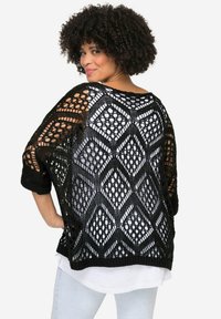 Schwarze Häkeljacke mit offenen Diamantmustern, Dreiviertelärmeln und einem strukturierten, leichten Design, getragen über einem weißen Oberteil.