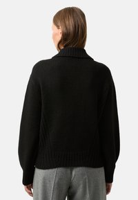 Black knitted sweater with a high collar, wide sleeves, and textured patterns at the back; paired with grey, pleated trousers.