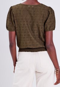 Blouse courte vert olive à manches bouffantes, motif tricot texturé, taille froncée et détails ajourés. Associée à un jean de couleur claire.