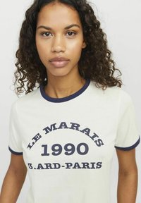 Crème t-shirt met korte mouwen en marineblauwe ringer details. De voorkant heeft gedurfde tekst: "LE MARAIS 1990 3.ARD-PARIS." Zachte katoenen stof.