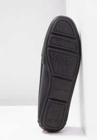 Chaussure en cuir noir avec une semelle en caoutchouc texturée présentant une bande de roulement à motifs. La semelle porte le logo "Calvin Klein".