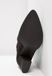 Zapato de tacón negro con punta afilada, textura de ante suave y suela de goma con ranuras horizontales para mayor agarre.