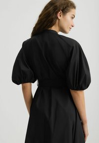 Käänatud naine lainetehiste juustega, kes kannab musta kleiti puff-sleeved varrukatega, pöörab veidi, näidates oma riietuse tagumist osa heleda tausta ees.