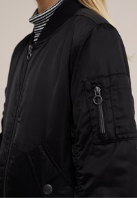 Close-up of a person wearing a black bomber jacket with a zipper and snap button pocket, over a black and white striped turtleneck.