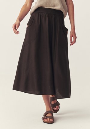 Kobieta ubrana w długą czarną spódnicę z przednimi kieszeniami i brązowe sandały z klamrą, stojąca na jednolitym jasnym tle.