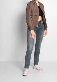 Chaqueta bomber marrón con acabado suave, puños acanalados y bolsillos frontales, combinada con jeans skinny azul deslavado y zapatillas blancas.