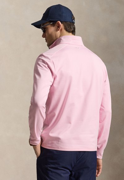 Άντρας που φοράει ανοιχτό ροζ μακρυμάνικο πουκάμισο, σκούρο μπλε παντελόνι, σκούφο σε σκούρη μπλε απόχρωση και γυαλιά ηλίου, στραμμένος με την πλάτη, με τα χέρια στις τσέπες.