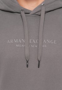 Pilkos spalvos džemperis su storomis raištelėmis ir užrašu "ARMANI EXCHANGE MILAN / NEW YORK" spausdintu per krūtinę derančios pilkos spalvos raidėmis.