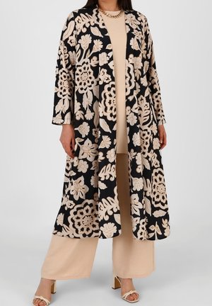 Femme portant un pantalon beige à jambes larges et un haut assorti à un long manteau noir à motif floral et un collier chaîne, debout les mains détendues.