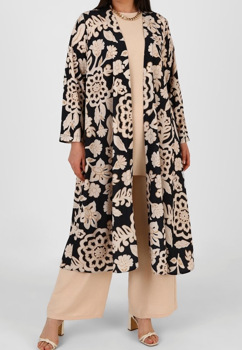 Femme portant un pantalon beige à jambes larges et un haut assorti à un long manteau noir à motif floral et un collier chaîne, debout les mains détendues.