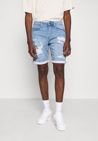 Pantalones cortos de mezclilla azul claro con detalles desgastados, puños enrollados, diseño de cinco bolsillos y cierre de botón, combinados con una camiseta beige y zapatillas blancas.